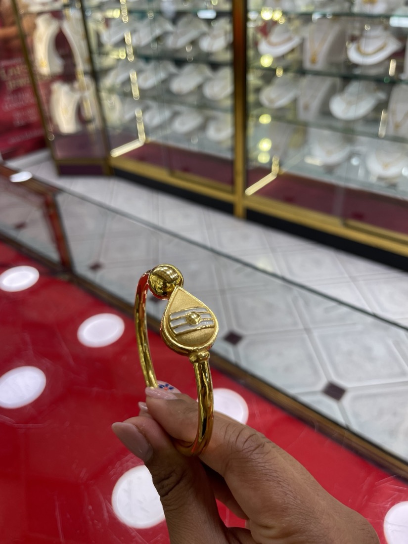 A gold Trishul bangle held up inside Lakshmis Jewellers, glass display cabinets of gold behind